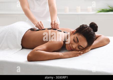 Schwarze Frau mit geschlossenen Augen und Kopf auf einem Handtuch liegend mit dem Gesicht nach unten auf dem Massagetisch, während die Therapeutin in der Kurklinik heiße Steine auf den Rücken legt Stockfoto