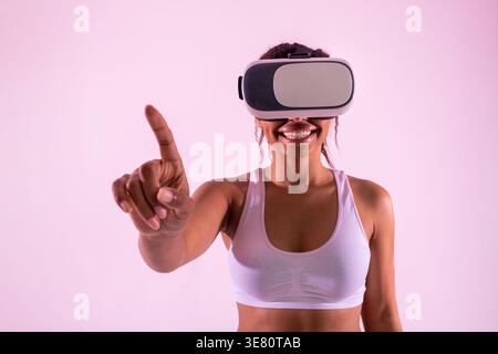 Schwarze Frau mit einer Virtual-Reality-Brille, die mit dem Finger zeigt, während sie im Studio vor violettem Hintergrund steht Stockfoto