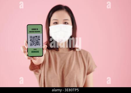 Asiatische Frau in medizinischer Maske zeigt Smartphone mit QR-Code nach dem Schuss des Impfstoffs gegen Coronavirus beim Stehen gegen rosa Hintergrund... Stockfoto