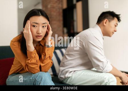 Asiatische Frau berührte ihre Schläfe nach einem Streit mit dem Mann, der im Wohnzimmer zu Hause auf dem Sofa saß Stockfoto