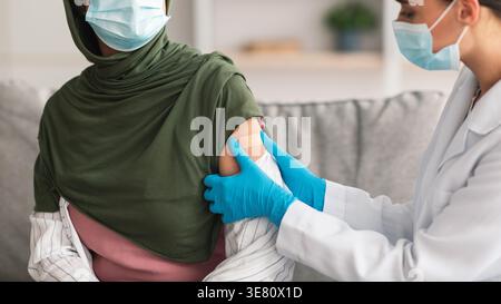 Zugeschnittenes Bild einer Ärztin aus dem Nahen Osten in Gesichtsmaske und Handschuhen, die Gipsverband auf den geimpften Arm einer muslimischen Frau mittleren Alters in Hij... Stockfoto