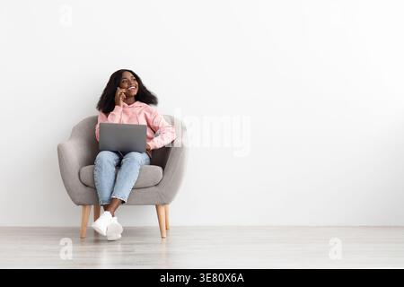 Lächelnde schwarze Frau in rosafarbener Jacke, die wegschaut und einen Anruf hat, sitzend auf einem Sessel mit Laptop vor der weißen Wand zu Hause Stockfoto