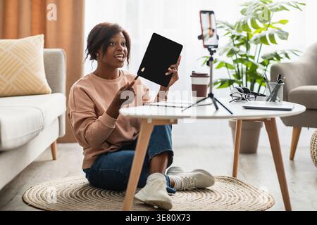 Schwarze Frau filmt sich auf einem Smartphone und zeigt ein Tablet mit leerem Bildschirm, das auf dem Teppich neben dem Tisch zu Hause sitzt Stockfoto