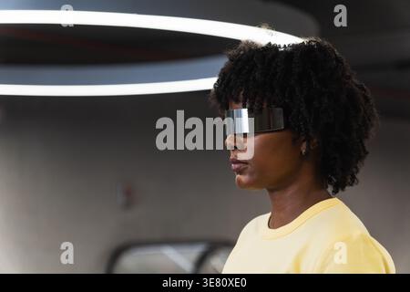 Schwarze Frau mit Afro, die eine futuristische, intelligente Brille trägt, die in einem Gebäude steht Stockfoto