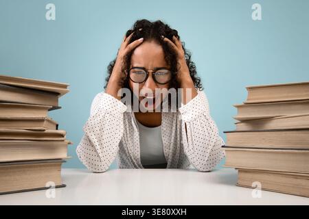 Gestresster schwarzer Teenager, der eine Brille trägt und Hände auf dem Kopf schreit, während er am Tisch sitzt, mit Büchern vor blauem Hintergrund in... Stockfoto