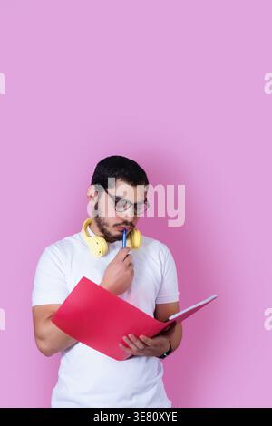 Arabischer männlicher Student mit Bart, Brille und Kopfhörer um den Hals, hält Stift und rosa Mappe und denkt, während er gegen rosa bac steht... Stockfoto