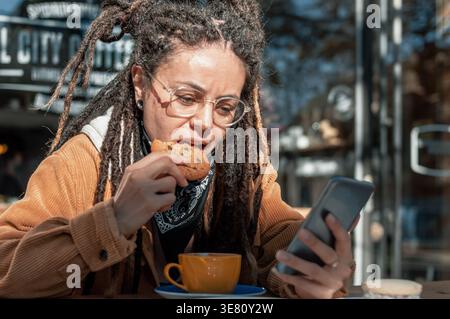 Hispanische Frau mit Dreadlocks, Brille und schwarzem Bandana am Hals mit Smartphone und Kekse beim Sitzen am Tisch mit Tasse... Stockfoto