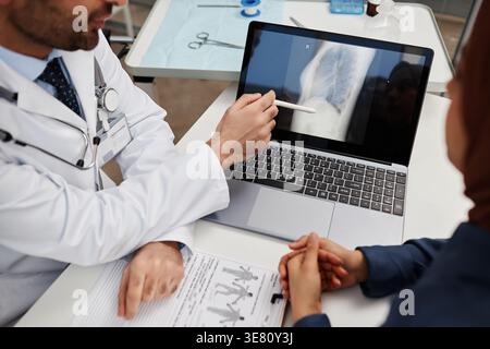 Zugeschnittenes Bild eines männlichen Arztes mit gemischter Rasse, der auf dem Laptop-Bildschirm auf das Röntgenergebnis zeigt, während er am Schreibtisch neben der Patientin in der Klinik sitzt Stockfoto