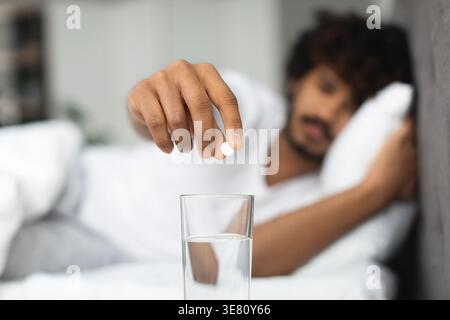Kranker asiatischer Mann mit lockigem Haar, der Pillen in ein Glas Wasser steckt, während er zu Hause auf dem Bett liegt Stockfoto