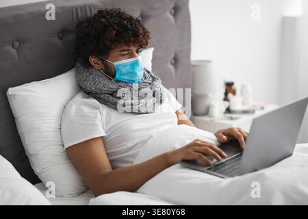 Kranker asiatischer Mann mit lockigem Haar, der Schal um den Hals und Gesichtsmaske mit Laptop trägt, während er zu Hause im Bett lag Stockfoto