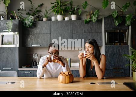 Schwarzes und gemischtes Paar trinkt Tee, während es am Tisch mit einer Schüssel Kekse in der Küche zu Hause sitzt Stockfoto