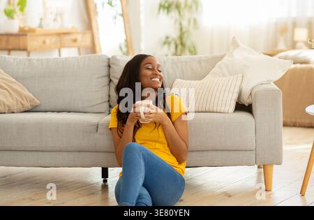 Fröhliche Schwarze Frau, die eine Kaffeetasse hält, während sie zu Hause im Wohnzimmer auf dem Boden sitzt Stockfoto