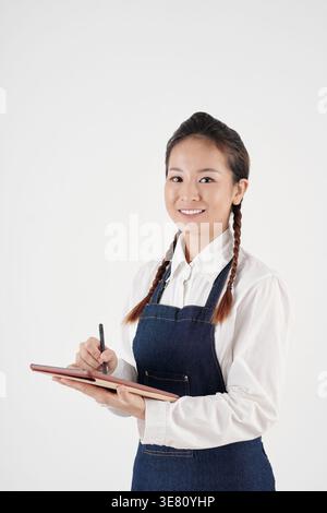 Asiatische Kellnerin mit Schürze auf Tablet, die vor weißem Hintergrund im Studio steht Stockfoto