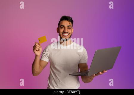 Arabischer Mann mit Bart, der Laptop und Kreditkarte hält, steht vor rosa und lila Farbverlauf Hintergrund im Studio Stockfoto