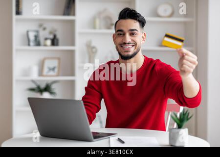 Arabischer Mann, der seine Kreditkarte zeigt und zu Hause mit Laptop sitzt Stockfoto