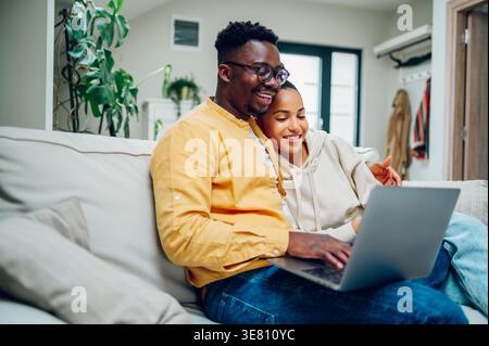 Schwarzer Mann, der seinen Laptop benutzt, während er auf dem Sofa neben einer gemischten Frau zu Hause sitzt Stockfoto