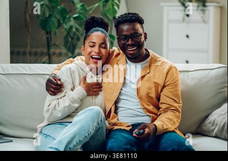 Lächelnder Schwarzer Mann und gemischte Frau mit einer Tasse Kaffee, die Spaß haben, zusammen auf dem Sofa im Wohnzimmer zu Hause zu sitzen Stockfoto