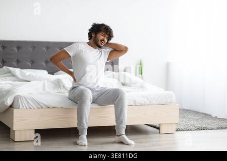 Bärtiger indischer Mann mit lockigen Haaren, der unter Rückenschmerzen leidet und im Schlafzimmer zu Hause auf dem Bett sitzt Stockfoto