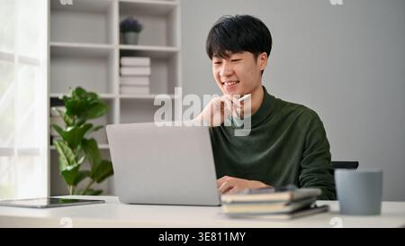 Asiatischer Mann, der am Laptop arbeitet, während er zu Hause am Tisch sitzt Stockfoto