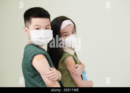 Asiatische Jungen und Mädchen mit medizinischen Masken mit Klebepflastern an Armen, die in der Klinik an der Wand stehen Stockfoto