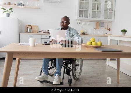 Behindertengerechter Geschäftsmann mit schwarzer Glatze im Rollstuhl mit Brille, Leser Dokumente am Tisch mit Laptop und Kaffeetasse in der Küche zu Hause Stockfoto