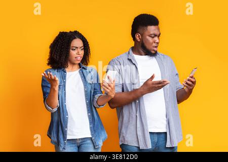 Schwarzes Paar, das mit schockierten Reaktionen auf ihr Smartphone schaut, während es vor gelbem Hintergrund steht Stockfoto