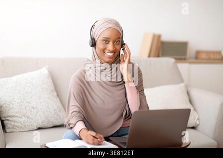 Schwarze muslimische Geschäftsfrau mit Hijab, die Notizen vor dem Laptop zu Hause schreibt Stockfoto