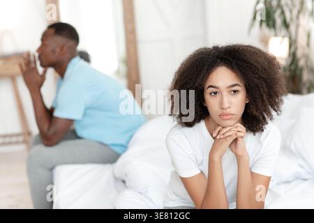 Schwarze Frau mit lockigen Haaren, die wegschaut und das Kinn auf den Händen anlehnt, während sie im Schlafzimmer zu Hause auf dem Bett gegen ihren Freund sitzt Stockfoto