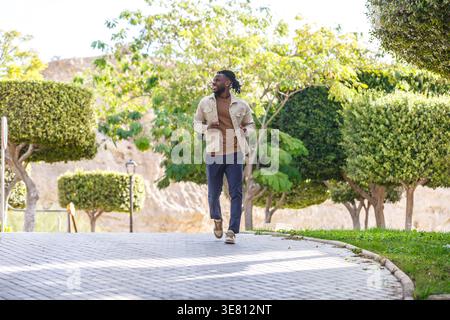 Schwarzer Mann mit Bart in beiger Jacke und blauer Hose, der am sonnigen Tag im Park wegschaut Stockfoto
