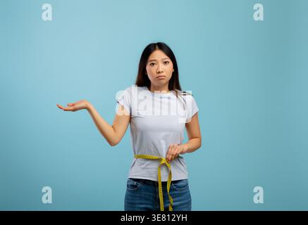Asiatische Frau, die ihre Taille mit Klebeband misst, während sie im Studio vor blauem Hintergrund steht Stockfoto