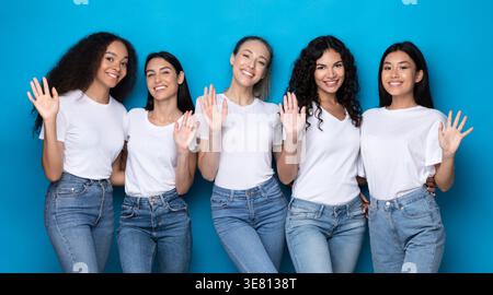 Eine Gruppe von Frauen in weißen Hemden und Jeans, die im Studio vor blauem Hintergrund die Hände winkten Stockfoto