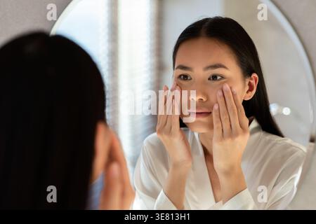 Asiatische Frau im Bademantel, die ihre Wangen berührt, während sie vor dem Spiegel im Badezimmer zu Hause steht Stockfoto