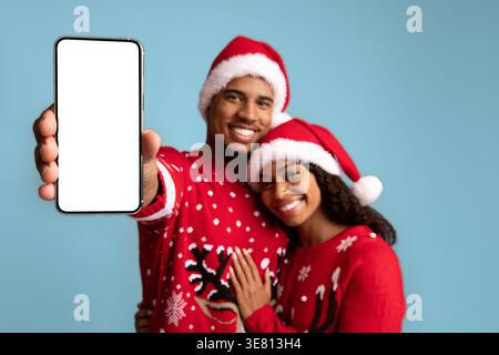 Lächelndes schwarzes Paar mit Weihnachtsmützen und Pullover, das Smartphone umschließt und zeigt, während es im Studio vor blauem Hintergrund steht Stockfoto