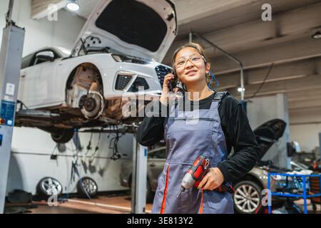 Chinesische Mechanikerin, die Overalls und Schutzbrille trägt, einen kabellosen Bohrer hält und einen Anruf hat, während sie wegblickt, während sie in der Nähe des Autos steht... Stockfoto
