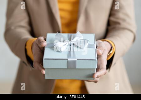 Zugeschnittenes Bild eines Mannes mit gemischter Rasse, der einen Mantel trägt und eine Geschenkbox hält, die im Studio vor grauem Hintergrund steht Stockfoto