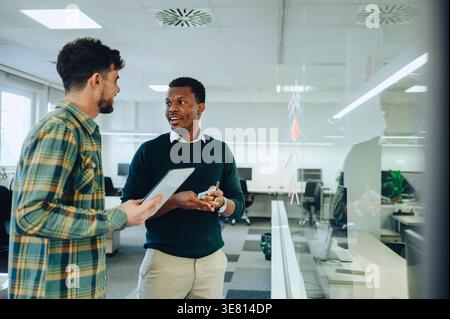Schwarzer Geschäftsmann, der seine Ideen auf Haftnotizen schreibt und den Projektplan mit einem männlichen kaukasischen Kollegen bespricht, der ein Tablett hält... Stockfoto