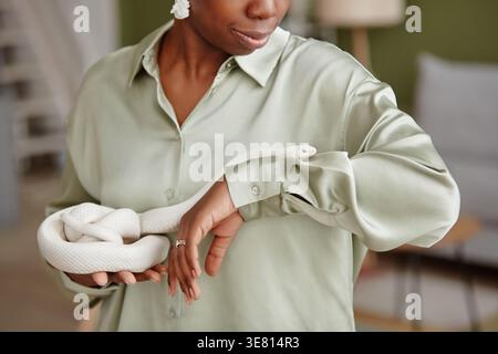 Abgeschnittenes Bild einer schwarzen Frau in Seidenbluse, die mit einer weißen Schlange über den Arm kriecht, während sie zu Hause steht Stockfoto