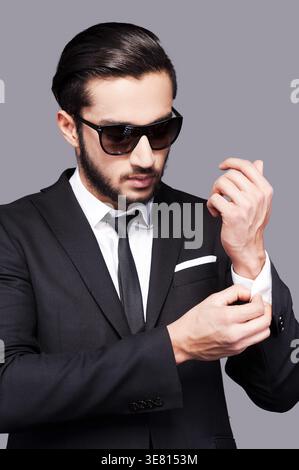 Geschäftsmann aus dem Nahen Osten, der eine Sonnenbrille trägt und seine Ärmel anpasst, während er im Studio vor grauem Hintergrund steht Stockfoto