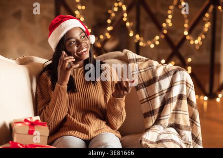 Schwarze Teenager-Frau, die Weihnachtsmann-Hut-Gesten-Hand trägt, während sie einen Anruf auf der Couch mit Geschenkboxen im Wohnzimmer zu Hause hat Stockfoto