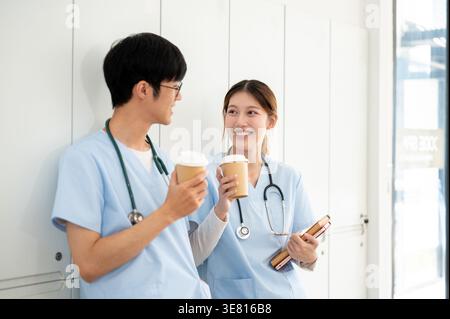 Asiatische männliche und weibliche Medizinstudenten, die Kaffee trinken und sich gegenseitig ansehen, während sie in der Umkleidekabine stehen Stockfoto