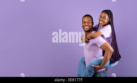 Fröhliche Schwarze Frau, die auf ihrem Mann steht, während sie im Studio vor violettem Hintergrund steht Stockfoto