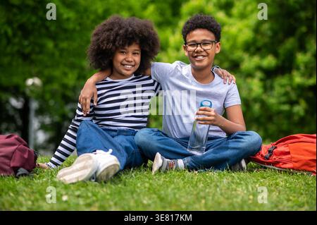 Schwarze Jugendliche umarmen sich gegenseitig, während sie im Park auf Gras sitzen Stockfoto