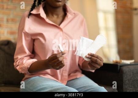 Zugeschnittenes Bild einer schwarzen Frau, die verschiedene Menstruationskissen hält, während sie zu Hause auf dem Sofa sitzt Stockfoto