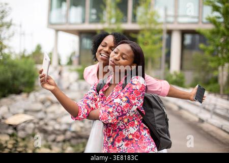 Afroamerikanische Teenagerin lehnt sich auf Metallgeländer und macht Selfie mit ihrer Mutter, während sie tagsüber mit dem Rucksack im Park steht Stockfoto