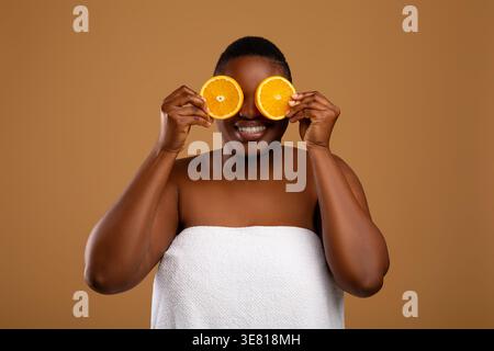 Schwarze Frau im Badetuch bedeckt Augen mit Zitrusfrüchten, die vor braunem Hintergrund im Studio stehen Stockfoto