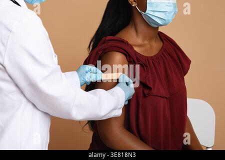 Schwarze Frau, die eine Gesichtsmaske trägt, sitzt auf einem Stuhl mit Schwester, die Gips auf ihren Arm vor beigem Hintergrund im Studio legt Stockfoto