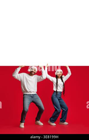 Lächelndes Paar aus dem Nahen Osten mit Weihnachtsmützen und Pullover, die großes weißes Brett halten, während sie im Studio vor rotem Hintergrund stehen Stockfoto