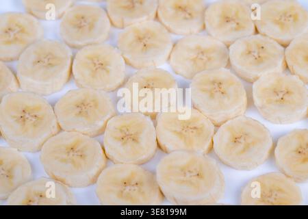In Scheiben geschnittene Bananen in weichem Gitter. Nahaufnahme reifer Bananenscheiben auf weißer Oberfläche, warmes direktionales Licht erzeugt sanfte Schatten, Betonung auf Creme Stockfoto