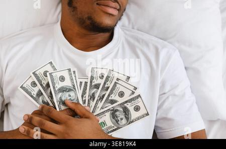 Schwarzer Mann in weißem Hemd, der Geld umarmt, während er zu Hause auf dem Bett schläft Stockfoto