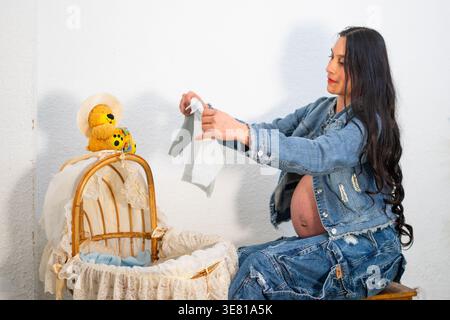 Hispanische schwangere Frau bereitet Babykleidung in der Wiege vor, während sie zu Hause auf dem Stuhl sitzt Stockfoto